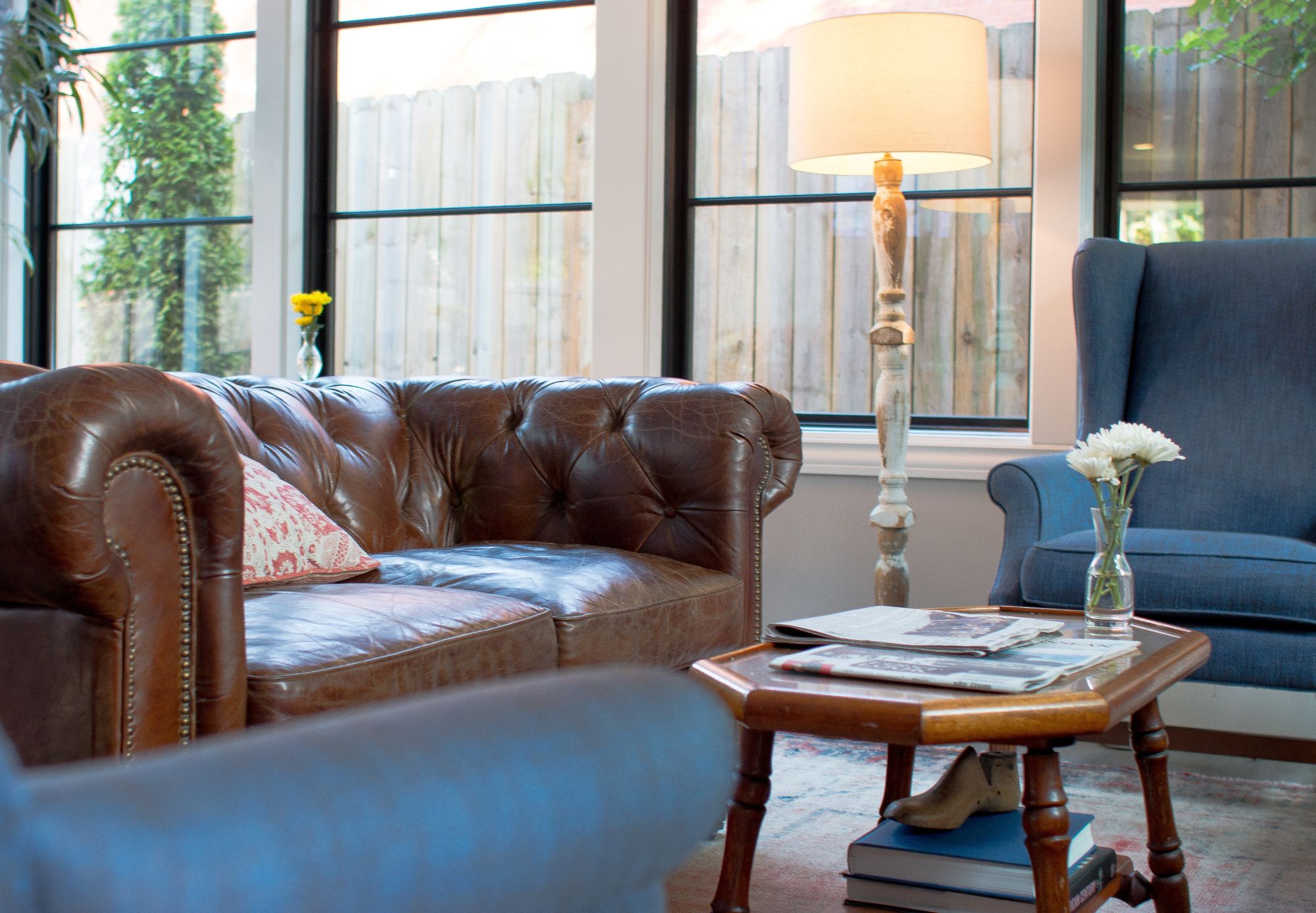 A cozy sunlit sitting room featuring a brown leather Chesterfield sofa, a blue armchair, and a wood coffee table